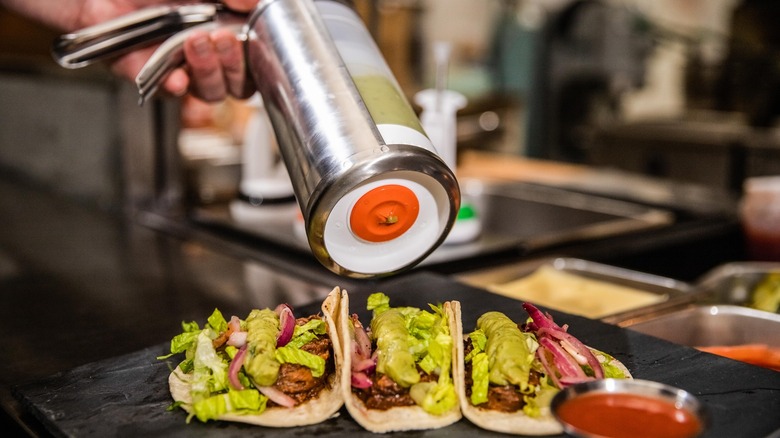 A food worker uses a sauce gun with a large valve to dress tacos with guacamole