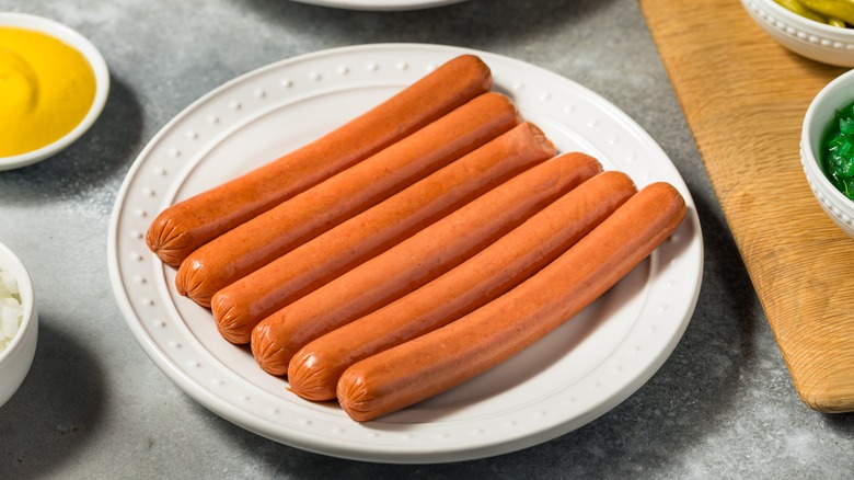 A plate full of uncooked hot dogs sits on a kitchen counter next to mustard dipping sauce and other toppings
