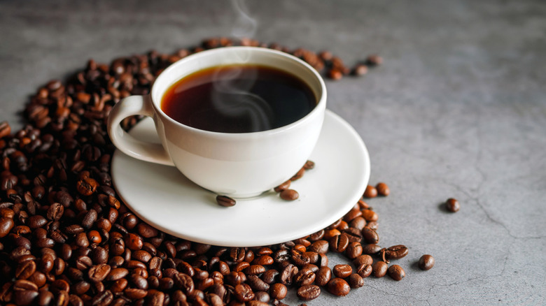 Steaming cup of coffee surrounded by coffee beans on a grey table.