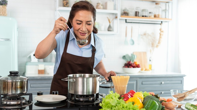 person happily cooking in a white kitchen, tasting soup from a ladle 