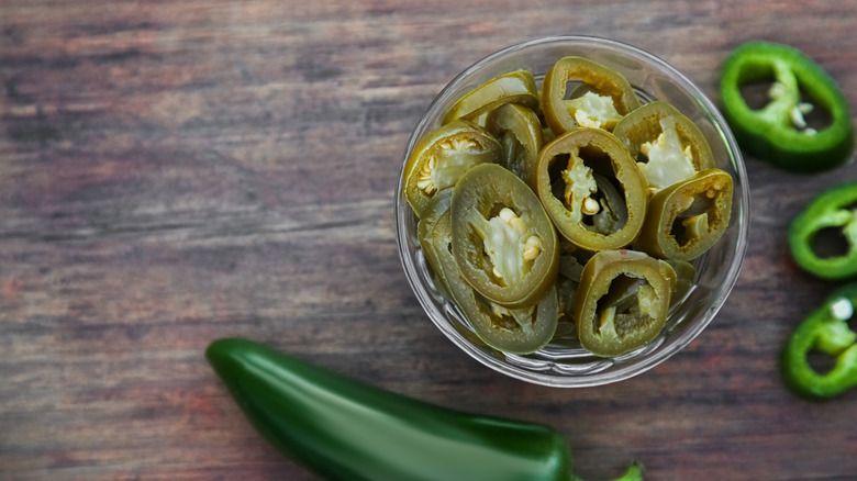 Pickled jalapenos sit in a small glass bowl with fresh jalapeno around it