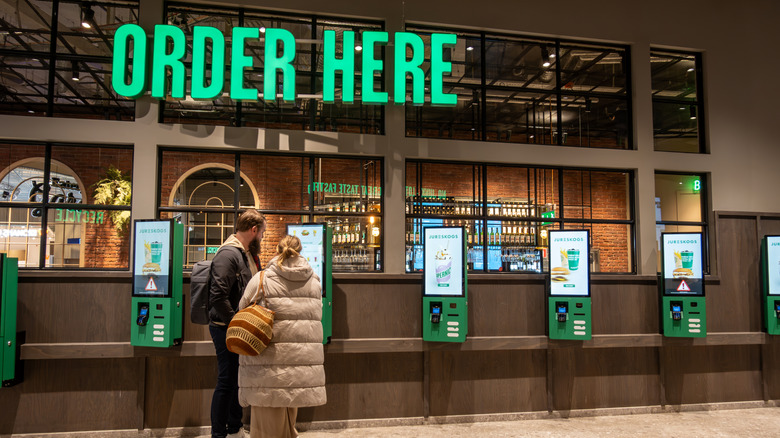A couple order at a self-service kiosk under a neon Order Here sign