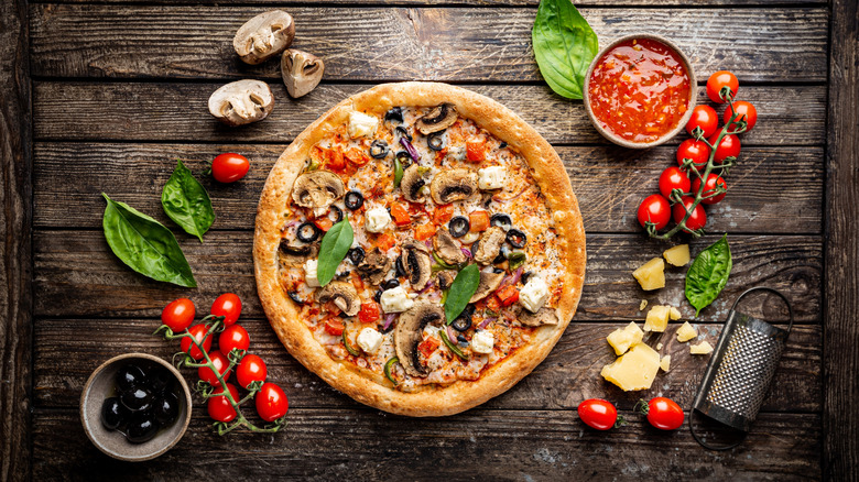 A veggie pizza surrounded by olives, cherry tomatoes, basil leaves, mushrooms, and cheese