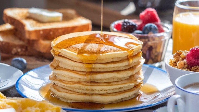 stack of pancakes with syrup drizzling down onto them, surrounded by toast, cereal, and beverages