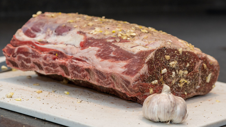 A whole raw prime rib roast, topped with garlic and black pepper, sits next to a bulb of garlic.