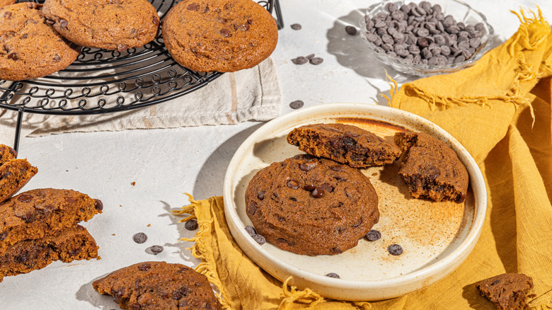 Pumpkin chocolate chip cookies on a plate and on a cooling rack with chocolate chips scattered around the table