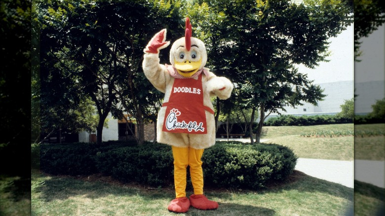Doodles the chicken, wearing an apron with his name and the Chick-fil-A logo, waves in an outdoor setting