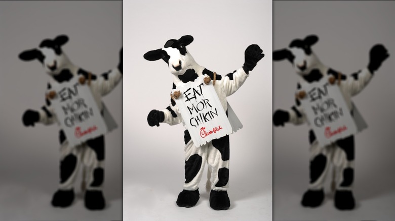 A person wearing a Chick-fil-A cow costume with a sandwich board that says "Eat Mor Chikin"