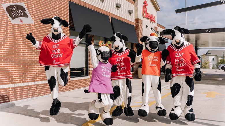 Five Chick-fil-A cow mascots jump in the air in front of a Chick-fil-A restaurant