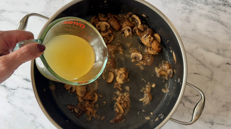 Hand holding cup of chicken broth over pan with mushrooms