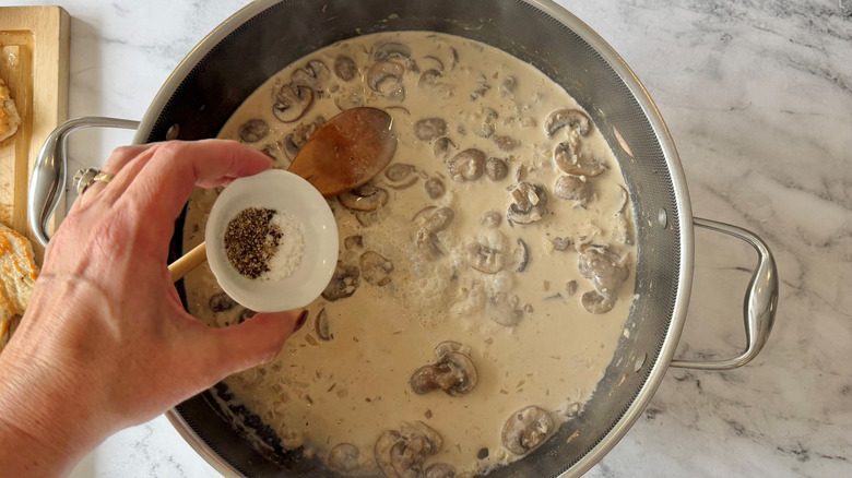 Hand holding bowl of salt and pepper over mushroom cream sauce in pan