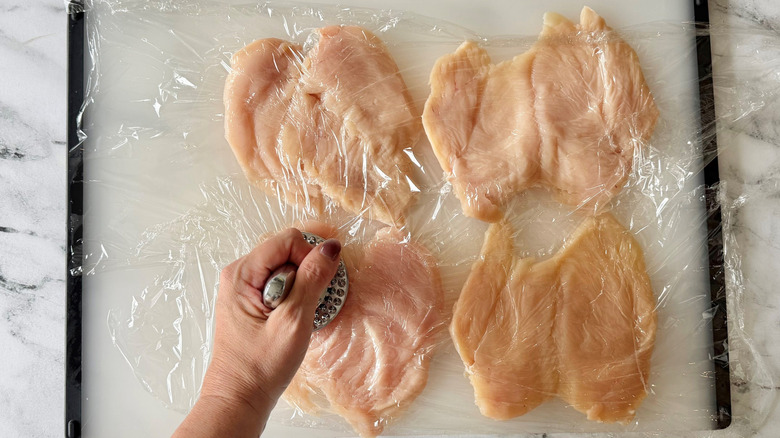 Hand using mallet to pound chicken breasts under plastic wrap