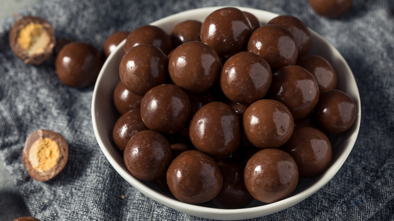 Chocolate-covered malted milk balls in a white ceramic bowl on a gray cloth surface, one of the chocolates is split open to show the malted milk interior