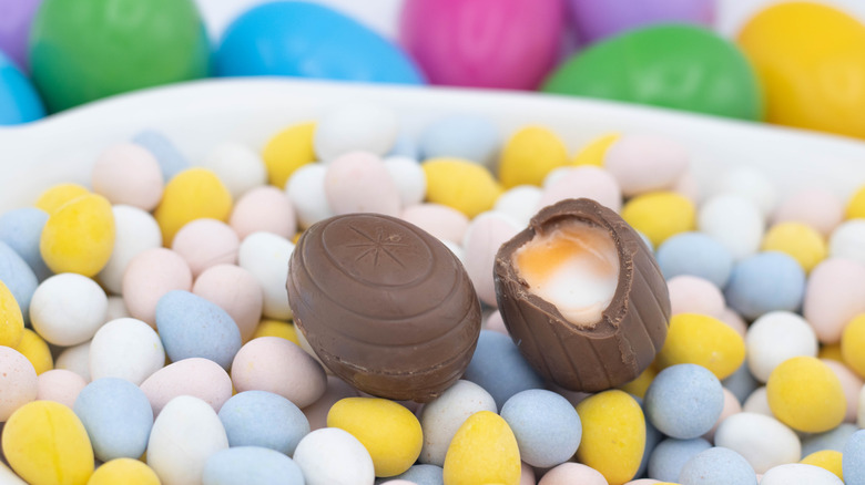 One intact Cadbury Creme Egg and one cracked open Cadbury Creme egg with its filling exposed, on mini candy-coated chocolate eggs in a white ceramic bowl with colorful plastic Easter eggs in the background