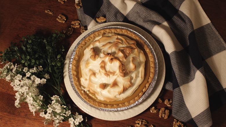 Toasty meringue tops a butterscotch pie served on with plate near shelled walnuts, a floral arrangement and blanket
