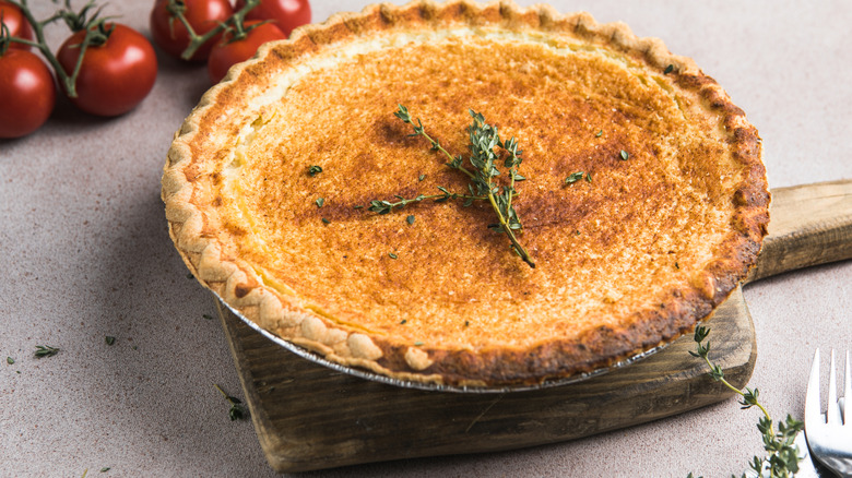 Chess pie topped with herbs atop a wooden tray in front of tomatoes on a vine