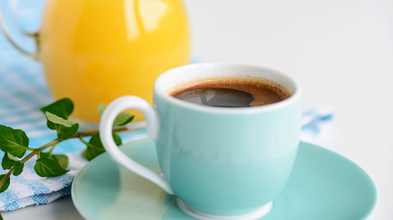 A cup of coffee on a saucer sits in front of a pitcher of orange juice.