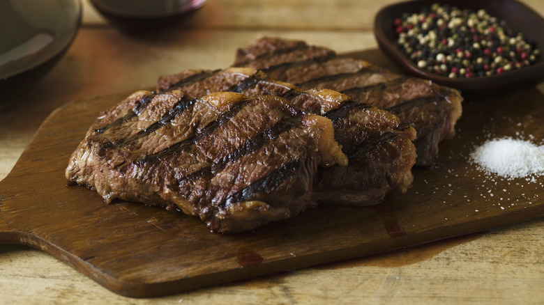Grilled steaks sit on a wooden board with salt and whole peppercorns