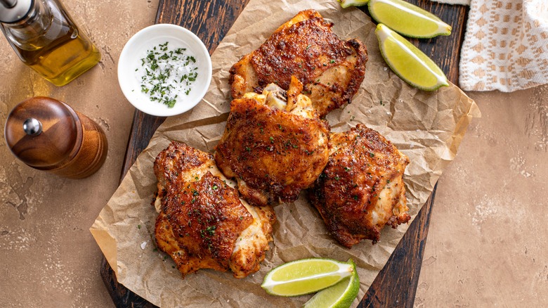 Baked chicken thighs, heavily seasoned, rest on a piece of parchment paper with limes