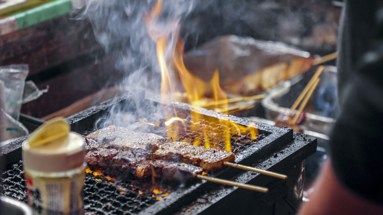 Skewered meat smoking on a fiery, charred grilltop