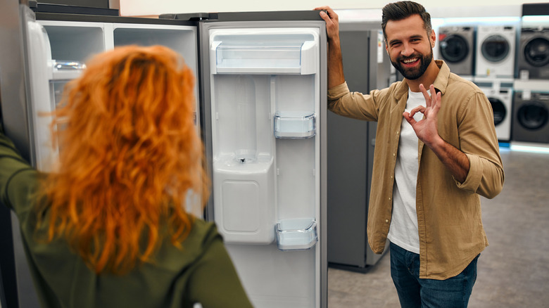 A couple stand near an open refrigerator in an appliance section of a store.