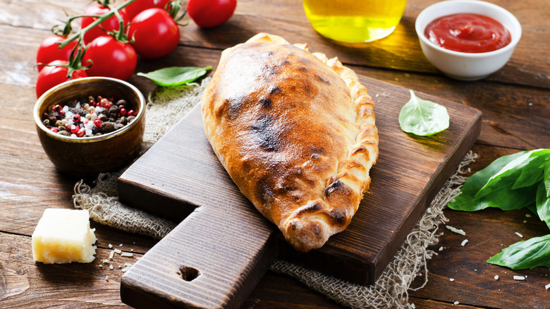 A calzone sits on a wooden cutting board, with a small hunk of cheese, basil leaves, oil, tomatoes, and ramekins of peppercorns and sauce sitting adjacent