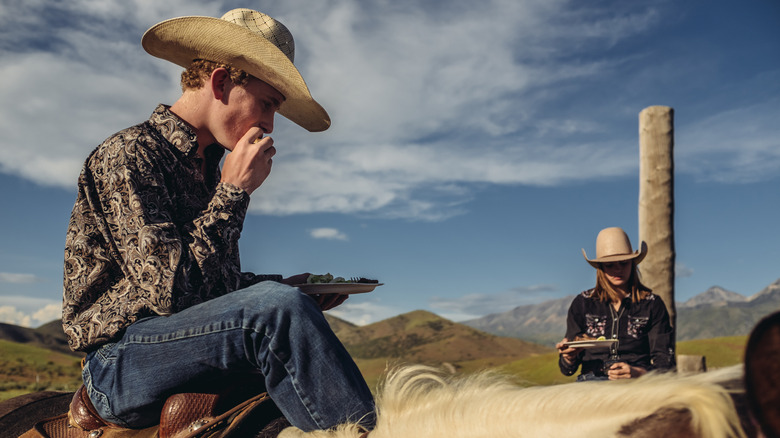 A cowboy eats lunch on his horse next to a cowgirl.