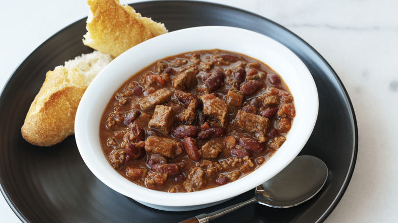 A white bowl of chile con carne sits on a black plate with bread and a spoon.