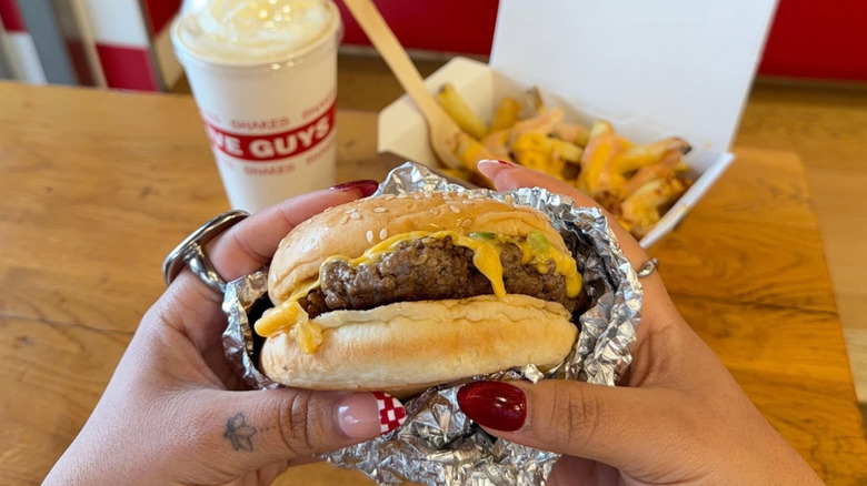 Someone holds a Five Guys burger, with fries and a shake on the table.