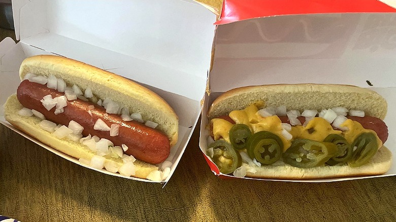 Two hot dogs with various garnishes sit in to-go boxes at 7-Eleven