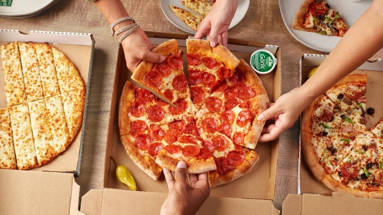 Several Papa John's pizzas and cheese sticks sit on a wooden table as people take pieces