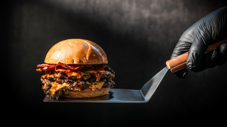 A person holds a gourmet bacon cheeseburger on a spatula against a black background
