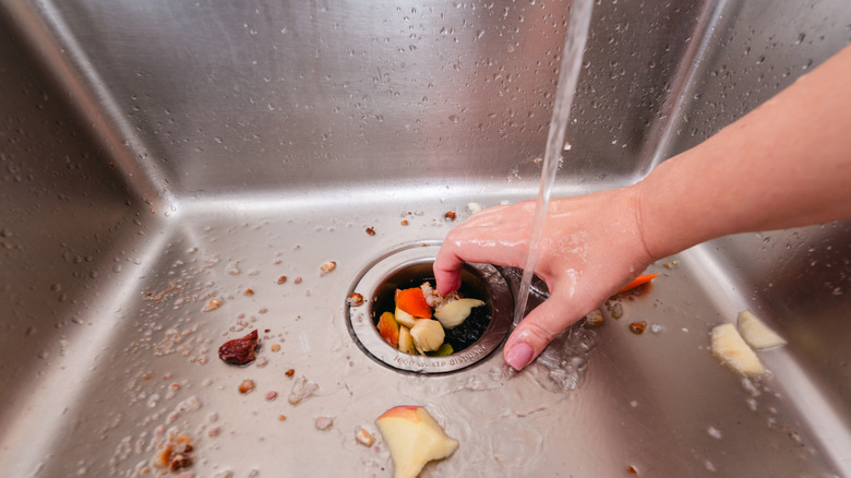 A hand pushes food into a garbage disposal in a stainless steel sink