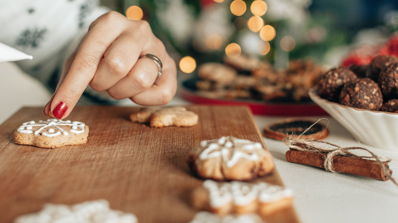 A hand decorating a cookie on a wooden board surrouunded by baked treats and cinnamon and a Christmas tree in the background