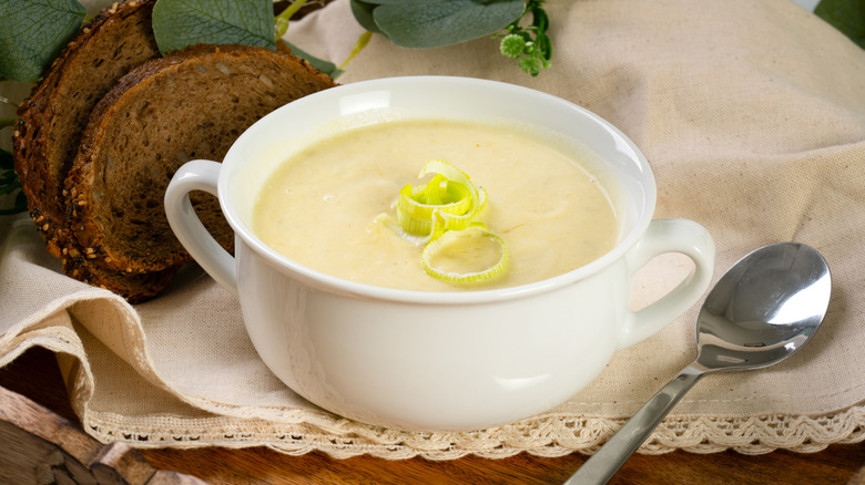 A simple bowl of potato soup, topped with leeks and sitting beside some dark bread.