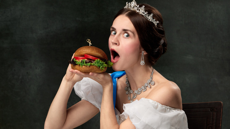 A woman wearing a crown, a white dress, and a blue royalty sash prepares to take a bite of a hamburger