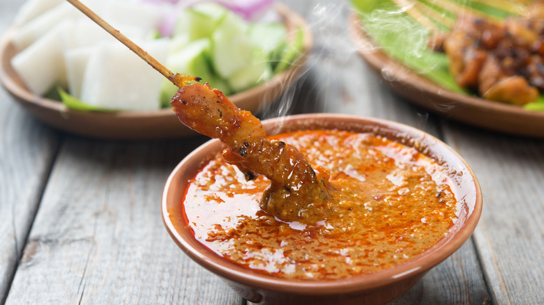 Cooked chicken on a skewer being dipped in steaming peanut sauce with vegetables and other meats blurred in the background, all on a light wooden table