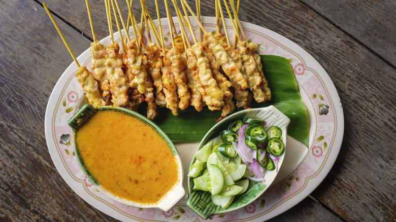 Multiple chicken skewers on a a pink and white plate with small bowl of peanut sauce and another small bowl of cucumbers, jalapenos, and onions, all on a wooden table