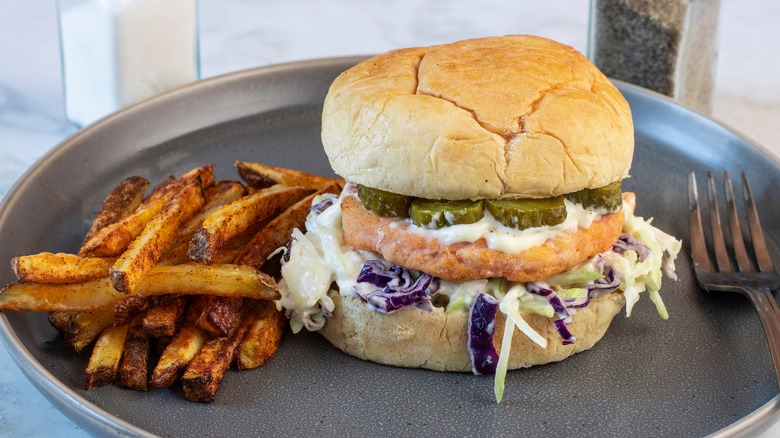 A salmon burger that has pickles and coleslaw, with a side of hand-cut french fries