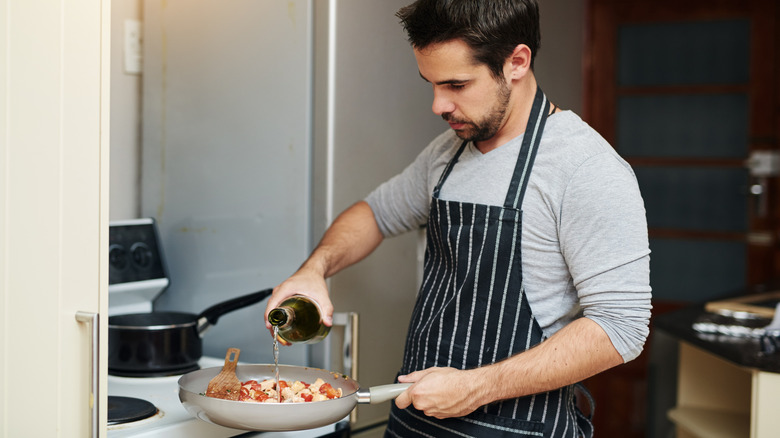 A man in an apron standing over the stovetop, pouring alcohol into his cooking