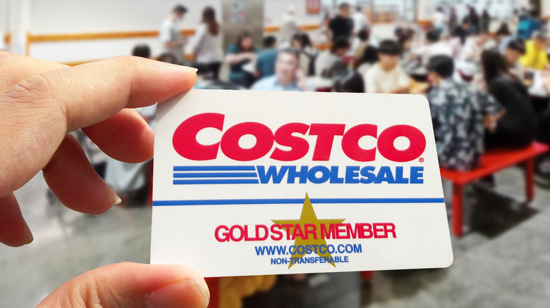 A hand holds up a Costco membership card with a Costco food court in the background