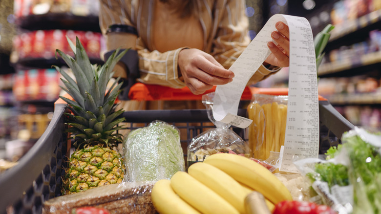 A woman's hands hold a long grocery receipt above a cart full of groceries