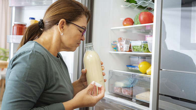 Woman standing at an open fridge smells the contents of a bottle to check for spoilage
