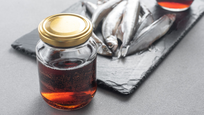 A glass jar of fish sauce with whole fish on a slate in the background