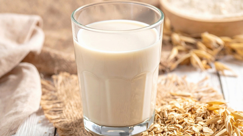 Oats with plants husk in the background next to a glass of oat milk.