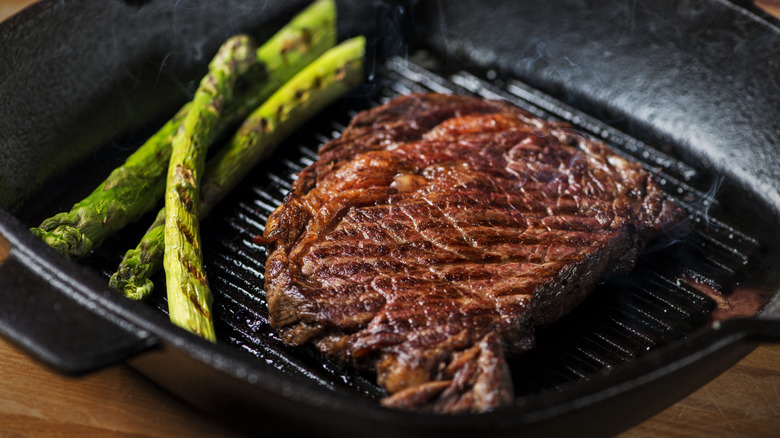 Ribeye steak being seared on a small grill beside asparagus