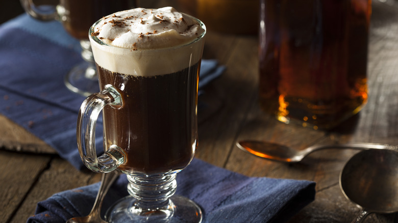 Irish coffee in a glass mug on a blue cloth napkin on a wooden table with a bottle of whiskey in the background