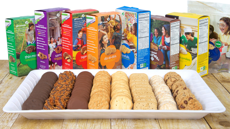 Various Girl Scouts cookies in single rows on a white tray with their boxes behind the tray