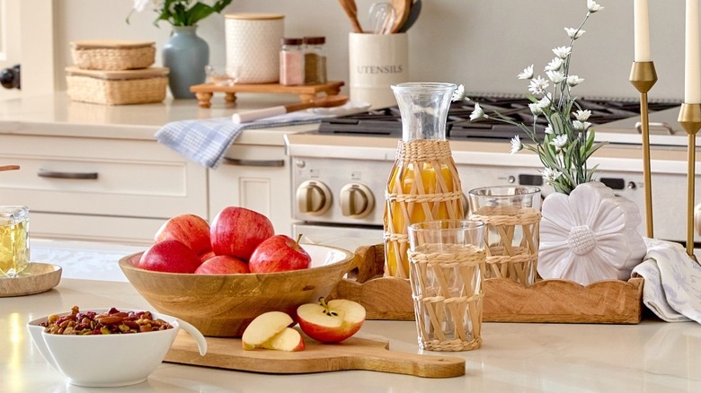 A rustic kitchen set sits on an island in a kitchen, featuring wooden bowls, cutting board, and tray with glasses and orange juice cannister wrapped in seagrass, and a vase with flowers