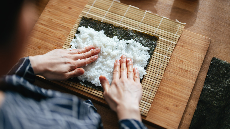 hands spreading sushi rice onto seaweed on bamboo mat